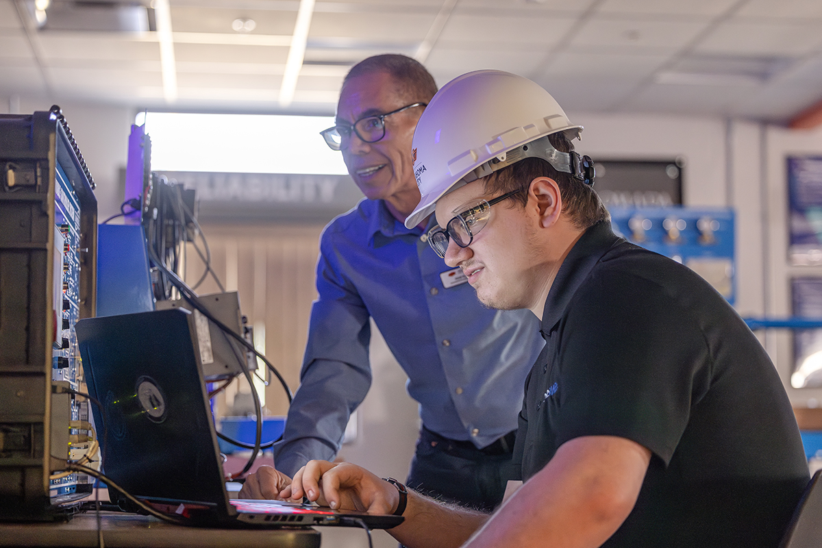 An instructor wearing a blue shirt stands beside a student wearing a white hard hat as they work together at a laptop connected to industrial networking and automation equipment in a technical training lab.