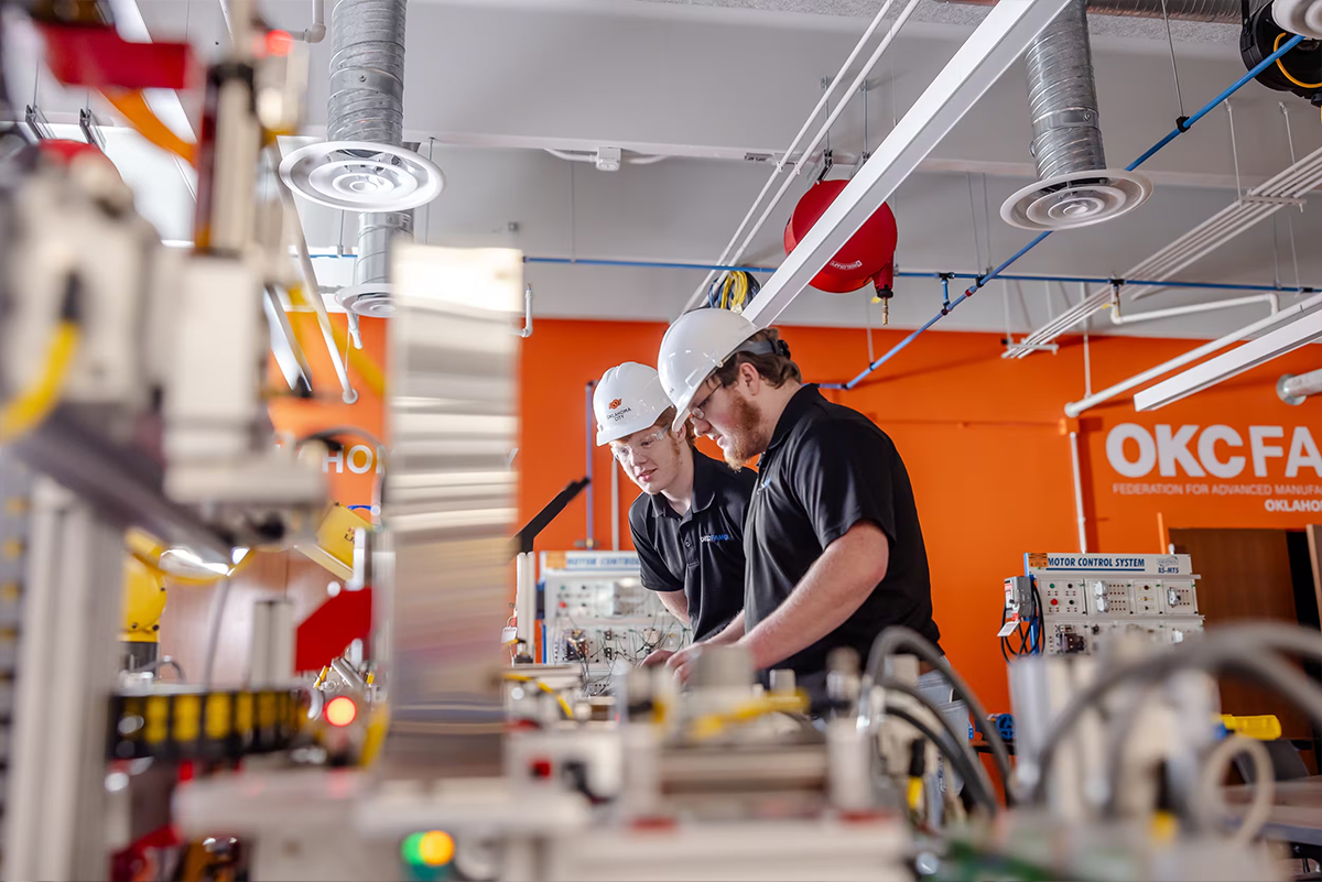 Two workers wearing hard hats inspect industrial automation equipment in a manufacturing lab.