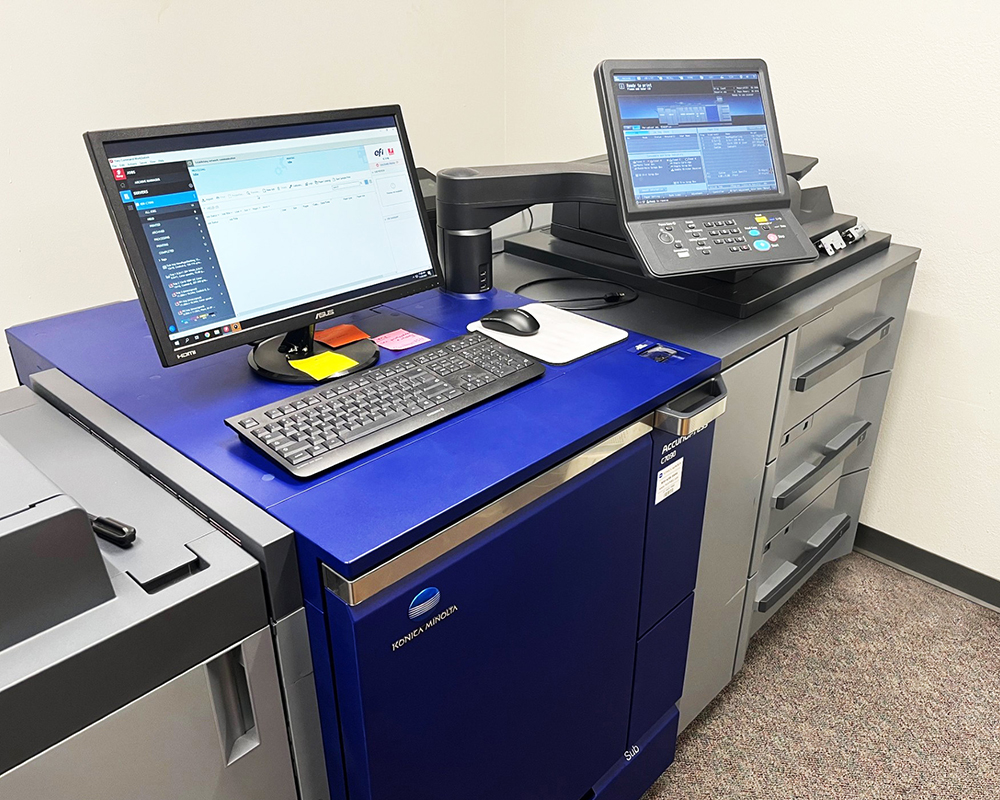 A modern large format Konica Minolta printer workstation with dual screens. The setup includes a keyboard and mouse, with a blue printer base.