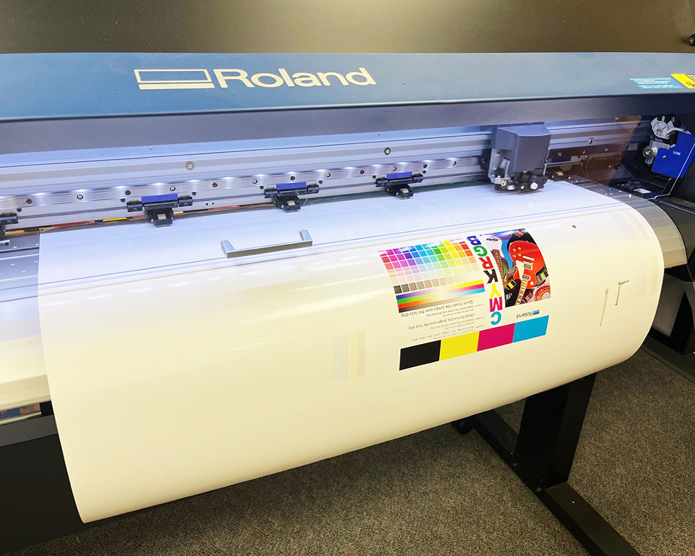 A large format Roland printer in operation, with a colorful test print featuring CMYK and RGB swatches emerging. The scene conveys a sense of precision and creativity.