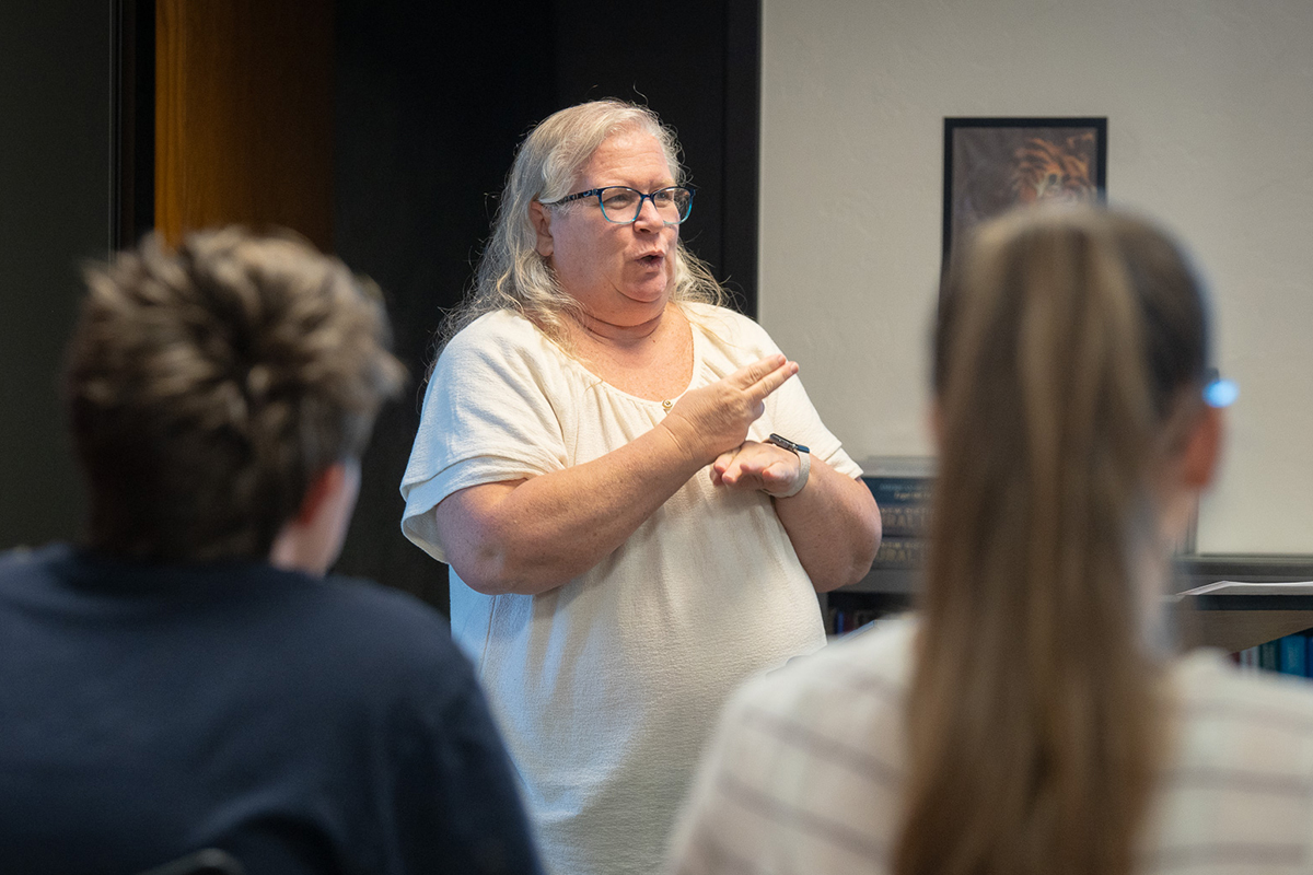A woman signs in front of an engaged audience. She is conveying focus and inclusivity in a classroom setting.