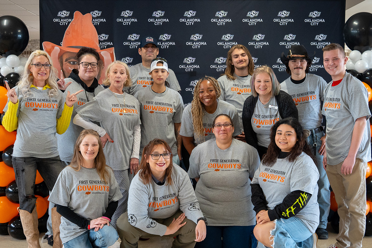 A group of students and staff wearing matching “First Generation Cowboys” T-shirts pose indoors in front of an Oklahoma State University–Oklahoma City backdrop with black and orange balloons.
