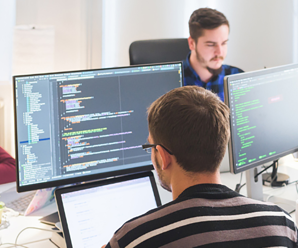 Two people sit across from each other in a computer lab environment, engaged in their work